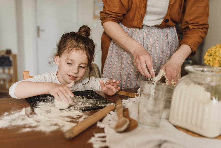 A young girl enjoys baking with an adult, kneading dough in a cozy kitchen.